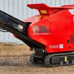 Photo of the Red Rhino 4000 Mini Crusher on some rough, sandy ground. This is a bright red piece of equipment, on black caterpillar tracks. A conveyor belt can be seen on the front of the mini crusher.