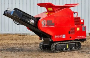 Photo of the Red Rhino 4000 Mini Crusher on some rough, sandy ground. This is a bright red piece of equipment, on black caterpillar tracks. A conveyor belt can be seen on the front of the mini crusher.