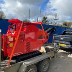 The bright red Rhino 4000 Mini Crusher tied on to a trailer and being towed to a site by a truck.