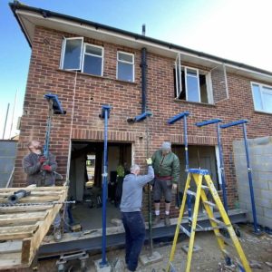 Photo of a partially complete red brick house in a construction site. Sticking out of the wall of the house are six Prop Pal systems. Three builders are using them to attach props to the ground.