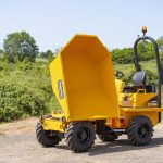 A photo of a bright yellow dumper on some rough ground with green hedgerows, trees and fields in the background. The dumper is branded Thwaites and has the large bucket tipped up and to the right of the photo.