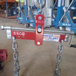 A photo of the Load Sling Adjuster in a work shop and hanging down from a large metal hook. The adjuster itself is bright red and has a Didcot Plant sticker on it. Metal chains are hanging down from the adjuster.